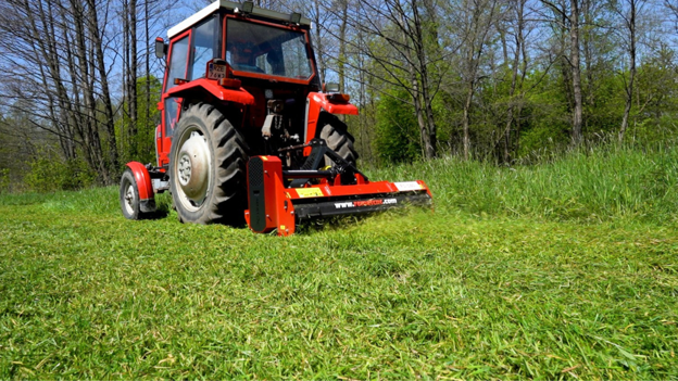 Flail mower attached to a tractor, cutting grass. A Remet FMS-140P 55" Flail Mower w/ Side Shift from Northern Woodsmen is attached to a tractor, cutting overgrown grass.