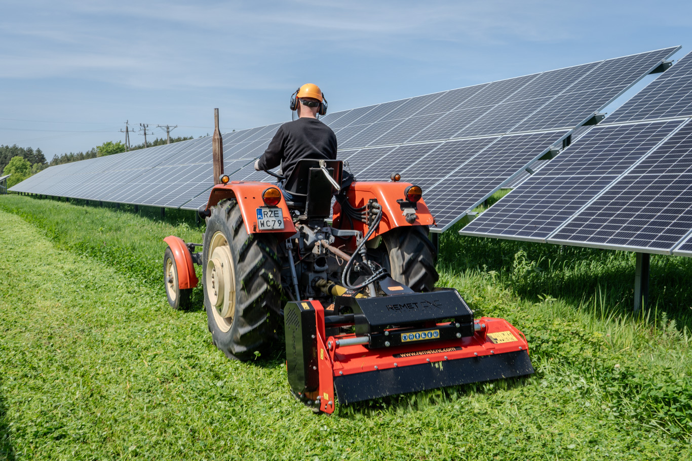 Original image of a tractor fitted with a red FMS-140P flail mower cutting grass along a solar panel field, showing the mower being used for controlled vegetation maintenance in a narrow working area.