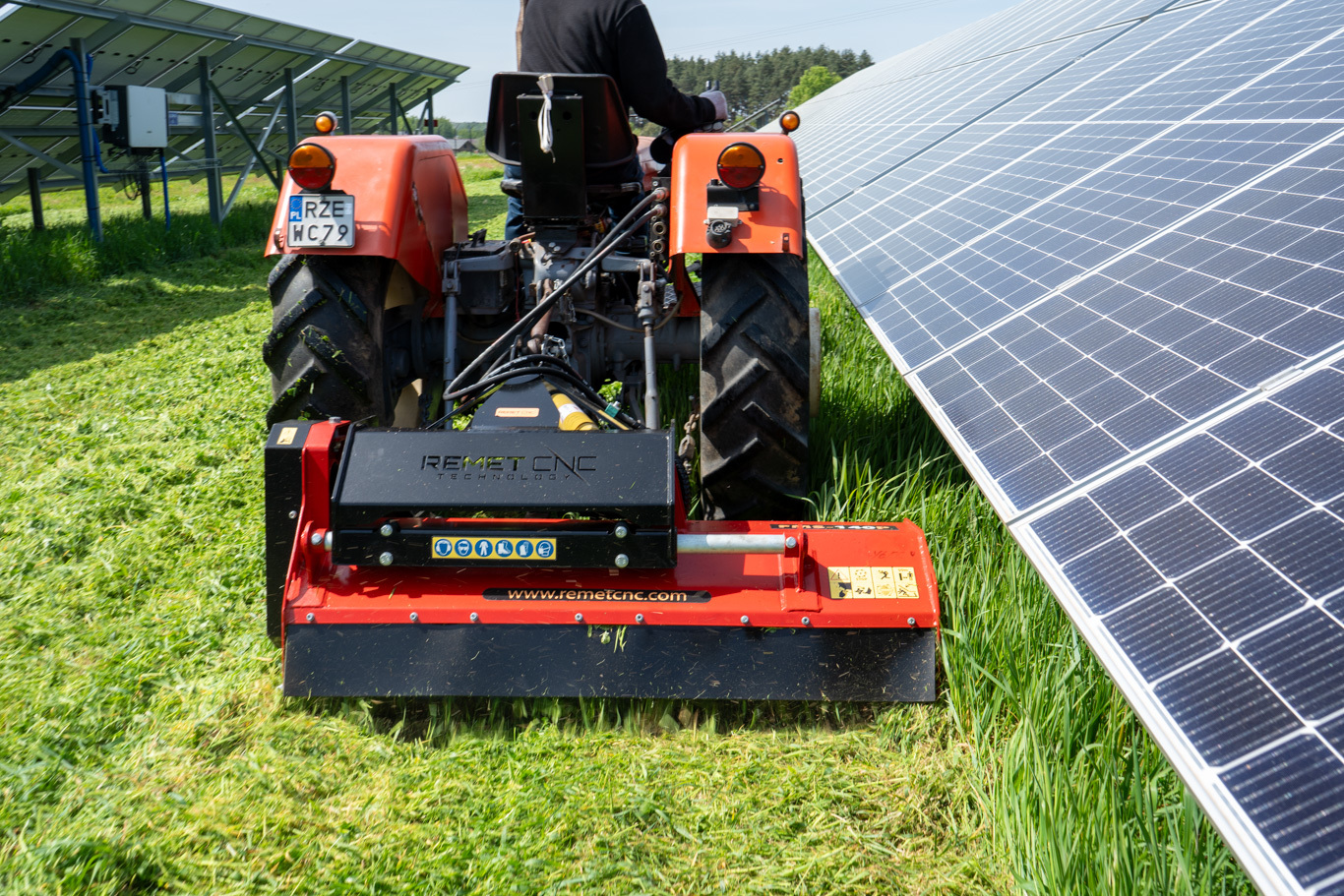 Original image of a tractor fitted with a red FMS-140P flail mower cutting grass in a narrow row between solar panels.