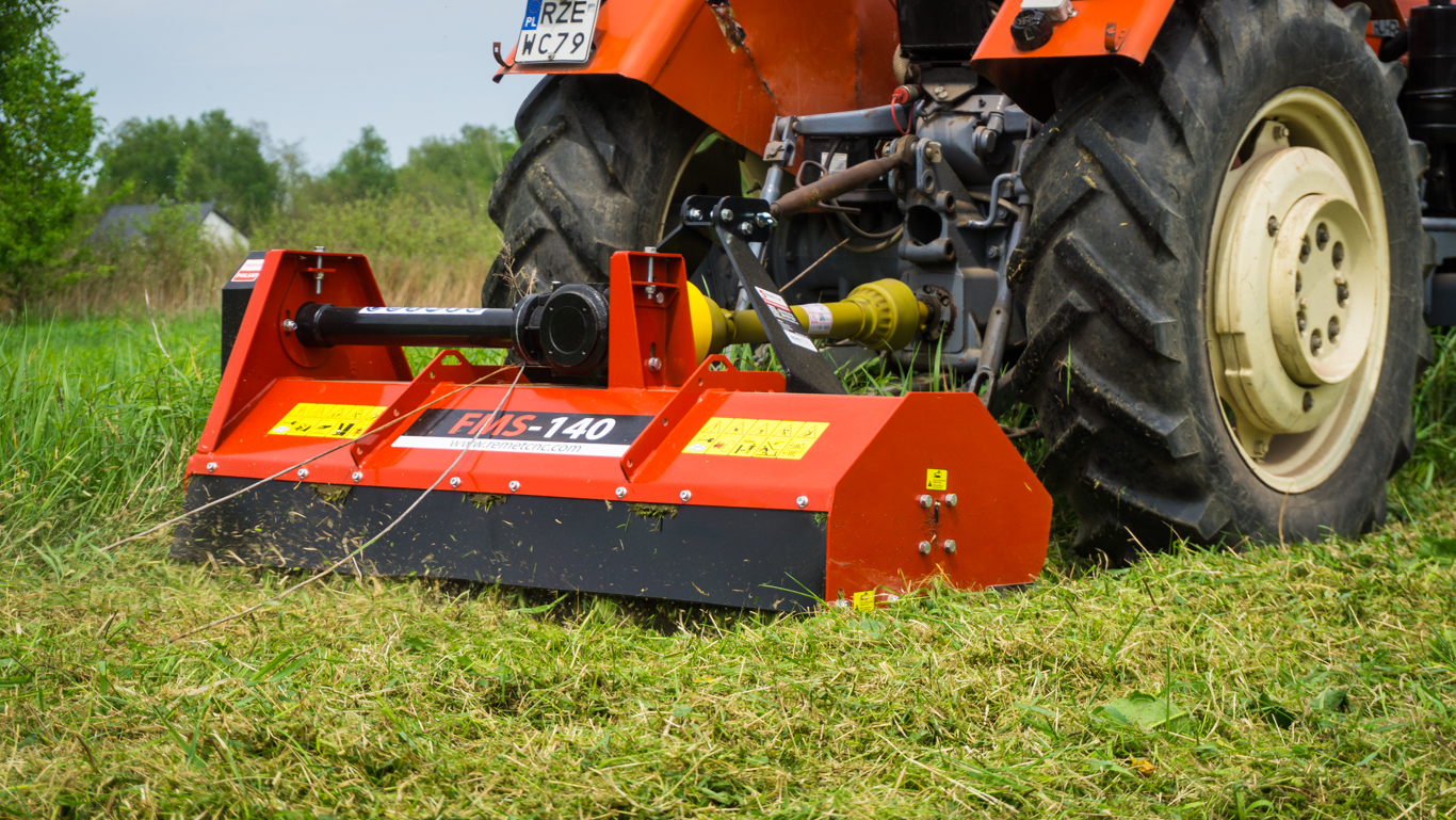 A red Remet FMS-140 55" Flail Mower from Northern Woodsmen is attached to a tractor, cutting and mulching overgrown grass.